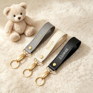 May include: Three leather-like wristlet keychains in gray, cream, and black, each featuring a gold-tone metal snap, a lobster clasp, and a circular ring. The gray strap is embossed with the name Aryah, the cream with Beren, and the black with Elowen.