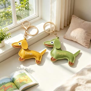 Puede incluir: Dos llaveros de perro salchicha. Uno es amarillo con el nombre "Ewan" y el otro es verde con el nombre "Peryn". Ambos tienen llaveros dorados y detalles marrones. Los llaveros están sobre una superficie blanca cerca de una ventana.