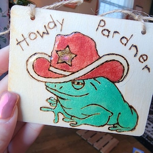 May include: Wooden sign with a green frog wearing a red cowboy hat with a gold star. The sign says "Howdy Pardner".