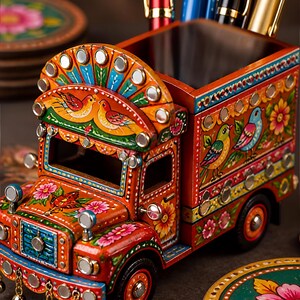Pu&ograve; includere: Un organizer da scrivania in legno a forma di camion, dipinto in arancione vivace con complessi motivi floreali e di uccelli. Il camion presenta piccoli specchi circolari, campanellini metallici pendenti e un vano cavo per riporre le penne.