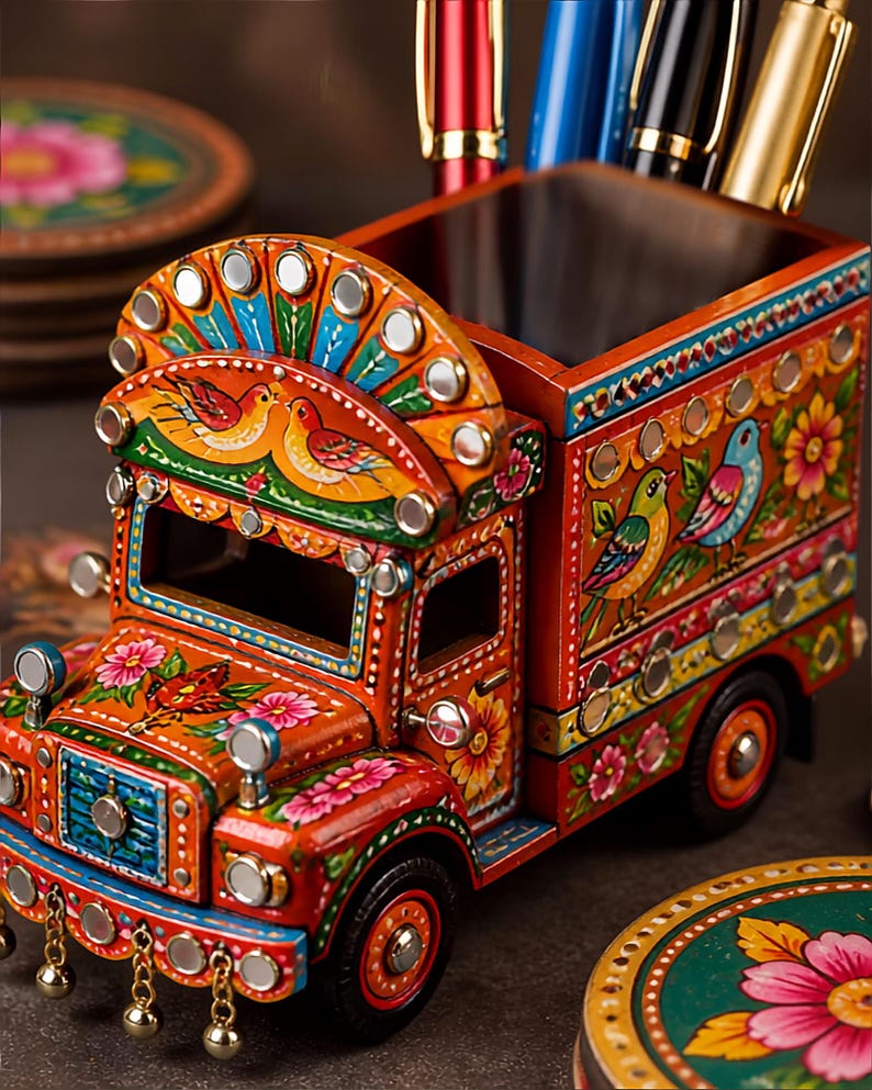 Pu&ograve; includere: Un organizer da scrivania in legno a forma di camion, dipinto in arancione vivace con complessi motivi floreali e di uccelli. Il camion presenta piccoli specchi circolari, campanellini metallici pendenti e un vano cavo per riporre le penne.