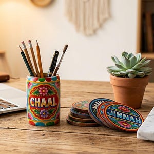 Pu&ograve; includere: Un portapenne cilindrico in legno dipinto con motivi floreali colorati e la parola CHAAL, contenente diverse matite e penne. Vicino si trovano sottobicchieri circolari in legno con disegni floreali e i nomi JINNAH e MASHA, oltre a una tazza.