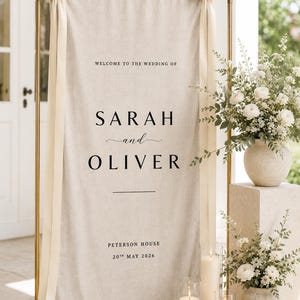 May include: A wedding welcome sign with the names "SARAH and OLIVER" in black text on a beige linen backdrop. The sign is held by a gold metal frame and adorned with cream-colored ribbons. The date is May 20th, 2026.