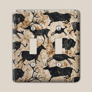 May include: A double light switch wall plate featuring a textured beige and brown background with a repeating pattern of black silhouettes of various animals, including deer and bison, resembling ancient cave paintings with four metal screws.