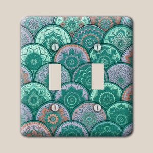 May include: A rectangular double light switch wall plate featuring a repeating pattern of overlapping circular mandala designs in shades of teal, mint green, lavender, and soft orange, with four silver screws securing the plate to the wall.