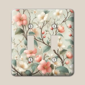 May include: A double light switch wall plate featuring a botanical pattern of pink and white cherry blossoms on thin brown branches with sage green leaves against a pale cream background, secured by four silver-colored metal screws.