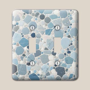 May include: A double light switch wall plate featuring a mosaic pattern of various sized stones in shades of light blue, dark blue, white, and grey. The rectangular cover has two vertical rectangular cutouts and four silver metal screws.