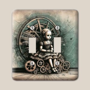 May include: A double light switch plate featuring a pale doll with large eyes sitting on a pile of bronze-coloured mechanical gears. Behind the doll is a large clockwork wheel set against a muted teal and beige background with thin lines.