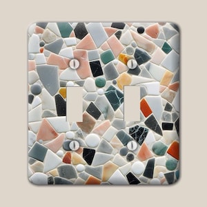 May include: A double light switch cover plate featuring a multi-coloured mosaic pattern of irregular stone-like fragments in shades of pink, grey, black, white, green, and orange, with four white screws positioned at the corners of the switch cutouts.