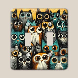 May include: A rectangular double light switch wall plate featuring a dense, colorful illustration of many cats with large, wide eyes. The cats are depicted in various shades of orange, blue, gray, and black, covering the entire surface area.