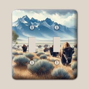 May include: A double light switch cover plate featuring a printed landscape of a herd of brown bison grazing in a dry, grassy field with sagebrush. In the background, tall, snow-capped blue mountains rise under a cloudy sky. Four screws are visible.