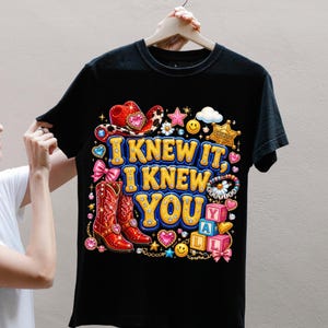 May include: A black t-shirt featuring a colorful graphic with the text "I KNEW IT, I KNEW YOU Y'ALL". The design includes a red cowboy hat, red cowboy boots, a sheriff badge, pink bows, smiley faces, stars, and alphabet blocks in a vibrant style.