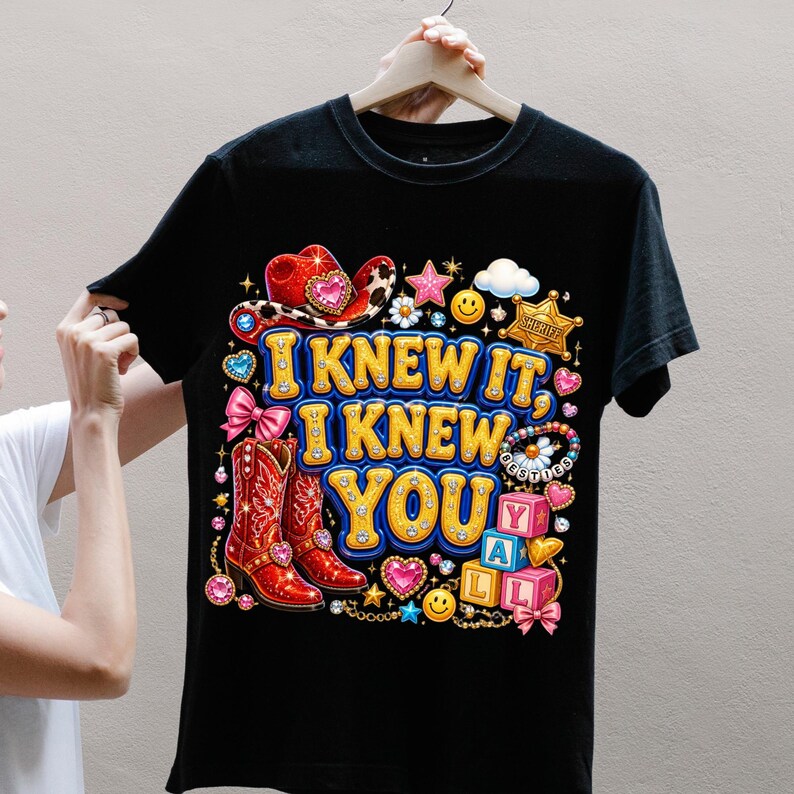 May include: A black t-shirt featuring a colorful graphic with the text "I KNEW IT, I KNEW YOU Y'ALL". The design includes a red cowboy hat, red cowboy boots, a sheriff badge, pink bows, smiley faces, stars, and alphabet blocks in a vibrant style.