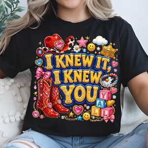 May include: A black t-shirt featuring a colorful graphic with the text I KNEW IT, I KNEW YOU. The design includes a red cowboy hat, red cowboy boots, a sheriff badge, pink bows, smiley faces, stars, hearts, and blocks spelling YALL.