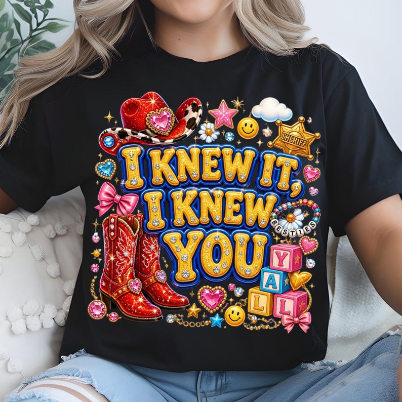 May include: A black t-shirt featuring a colorful graphic with the text I KNEW IT, I KNEW YOU. The design includes a red cowboy hat, red cowboy boots, a sheriff badge, pink bows, smiley faces, stars, hearts, and blocks spelling YALL.