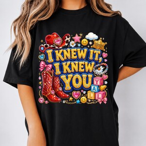 May include: A black t-shirt featuring a colorful graphic print with the text I KNEW IT, I KNEW YOU Y'ALL. The design includes red cowboy boots, a red cowboy hat, a sheriff badge, smiley faces, pink bows, stars, and alphabet blocks spelling Y'ALL.
