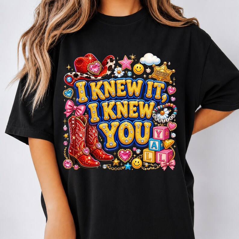 May include: A black t-shirt featuring a colorful graphic print with the text I KNEW IT, I KNEW YOU Y'ALL. The design includes red cowboy boots, a red cowboy hat, a sheriff badge, smiley faces, pink bows, stars, and alphabet blocks spelling Y'ALL.