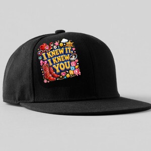 May include: A black baseball cap featuring a colorful graphic print on the front panel. The design includes the text I KNEW IT, I KNEW YOU, surrounded by illustrations of red cowboy boots, pink bows, stars, smiley faces, and alphabet blocks.