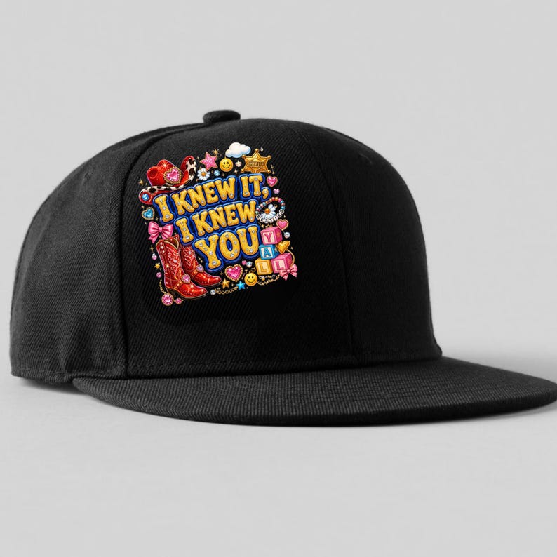 May include: A black baseball cap featuring a colorful graphic print on the front panel. The design includes the text I KNEW IT, I KNEW YOU, surrounded by illustrations of red cowboy boots, pink bows, stars, smiley faces, and alphabet blocks.