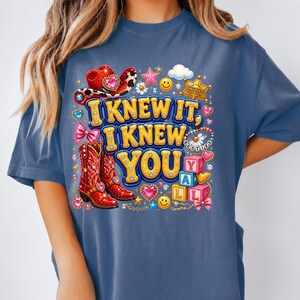May include: A blue t-shirt featuring a graphic print with the text I KNEW IT, I KNEW YOU. The design includes red cowboy boots, a red cowboy hat, a gold sheriff badge, pink bows, smiley faces, stars, hearts, and alphabet blocks spelling YALL.