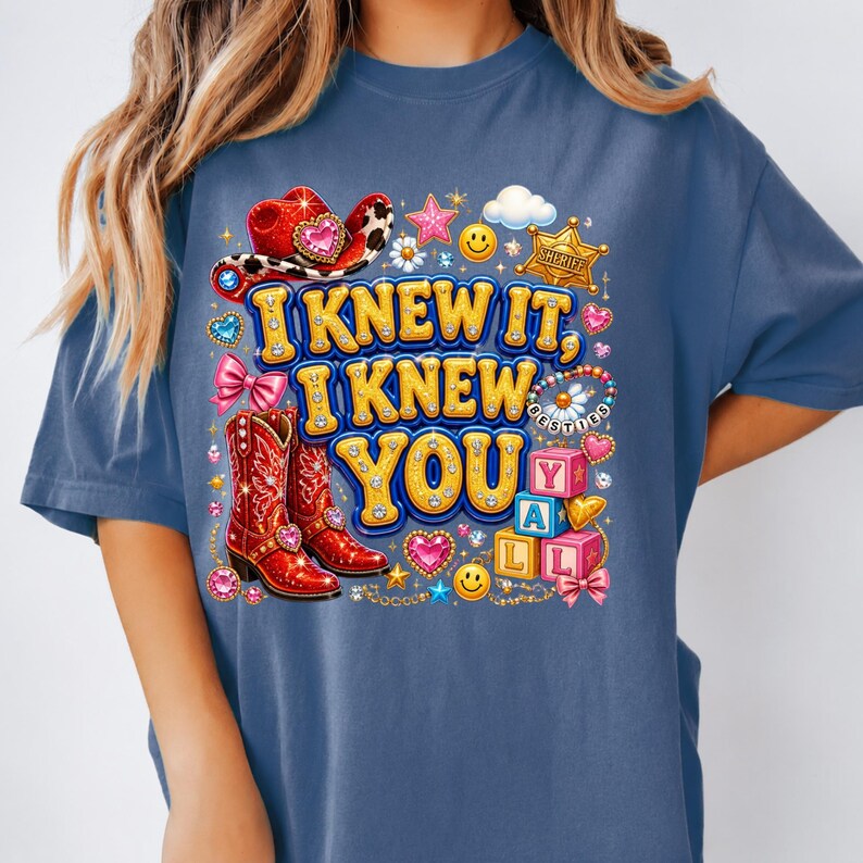 May include: A blue t-shirt featuring a graphic print with the text I KNEW IT, I KNEW YOU. The design includes red cowboy boots, a red cowboy hat, a gold sheriff badge, pink bows, smiley faces, stars, hearts, and alphabet blocks spelling YALL.