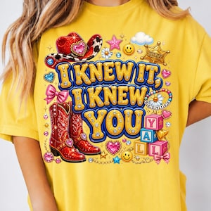 May include: A yellow t-shirt featuring a graphic print with the text I KNEW IT, I KNEW YOU. The design includes a red cowboy hat, red cowboy boots, a sheriff badge, smiley faces, pink bows, colorful hearts, stars, and stacked alphabet blocks.