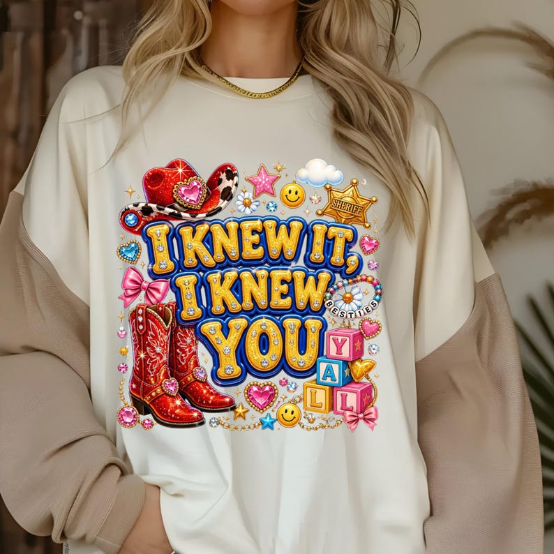 May include: A cream-colored sweatshirt featuring a graphic print with the text "I KNEW IT, I KNEW YOU". The design includes a red cowboy hat, red cowboy boots, a sheriff badge, pink bows, alphabet blocks, smiley faces, and various colorful hearts.