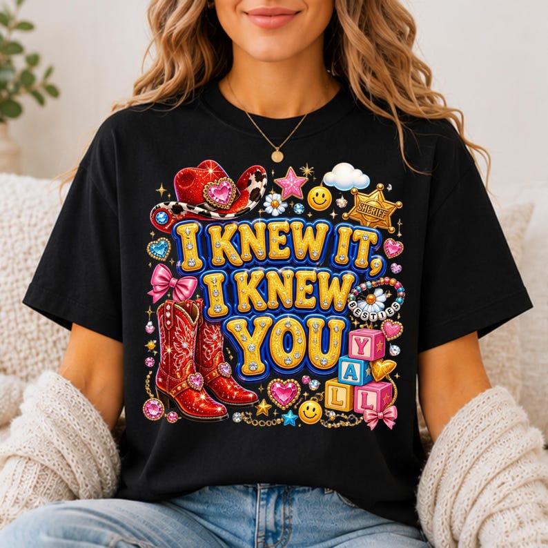 May include: A black t-shirt featuring a colorful graphic print with the text I KNEW IT, I KNEW YOU. The design includes red cowboy boots, a red cowboy hat, a sheriff badge, pink blocks spelling YALL, smiley faces, hearts, and stars.