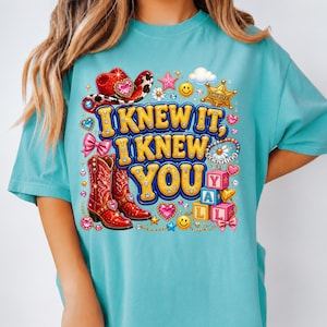 May include: A turquoise t-shirt featuring a graphic print with the text I KNEW IT, I KNEW YOU. The design includes a red cowboy hat, red cowboy boots, a sheriff badge, pink bows, smiley faces, hearts, stars, and alphabet blocks spelling YALL.