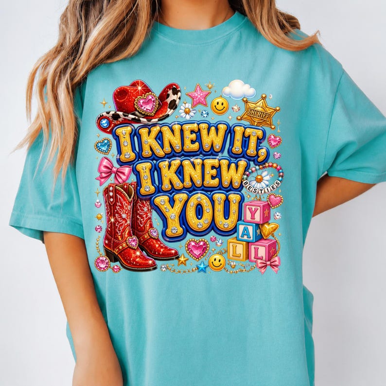 May include: A turquoise t-shirt featuring a graphic print with the text I KNEW IT, I KNEW YOU. The design includes a red cowboy hat, red cowboy boots, a sheriff badge, pink bows, smiley faces, hearts, stars, and alphabet blocks spelling YALL.