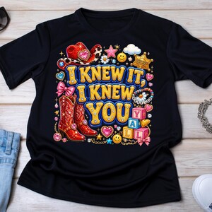 May include: A black t-shirt featuring a colorful graphic with the text I KNEW IT, I KNEW YOU. The design includes red cowboy boots, a red cowboy hat, a sheriff badge, pink bows, smiley faces, hearts, stars, and alphabet blocks spelling YALL.