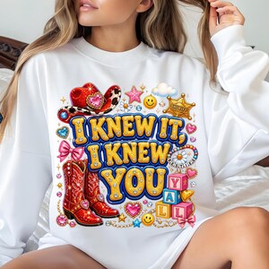 May include: A white long-sleeve sweatshirt featuring a colorful graphic print with the text I KNEW IT, I KNEW YOU. The design includes red cowboy boots, a red cowboy hat, a sheriff badge, pink bows, smiley faces, and stacked alphabet blocks.