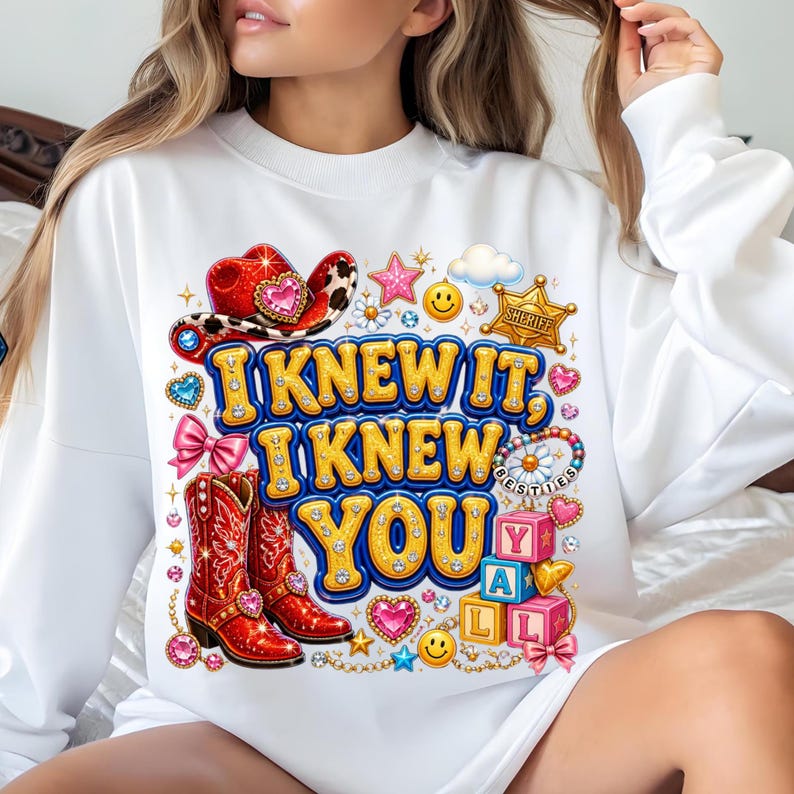 May include: A white long-sleeve sweatshirt featuring a colorful graphic print with the text I KNEW IT, I KNEW YOU. The design includes red cowboy boots, a red cowboy hat, a sheriff badge, pink bows, smiley faces, and stacked alphabet blocks.