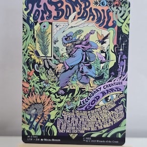 May include: A rectangular trading card titled Tom Bombadil featuring a colourful, psychedelic illustration of a bearded figure in a hat walking through a forest with sunflowers, a white bird, and purple mushrooms. The card includes game text.