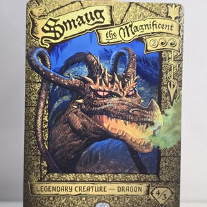 May include: A rectangular trading card featuring a gold-toned border with the text Smaug the Magnificent. The central illustration depicts a brown dragon with glowing pink eyes and smoke emitting from its mouth against a dark blue background.