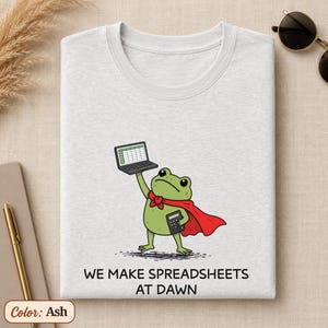 May include: An ash gray t-shirt featuring a graphic of a green frog wearing a red cape, holding a laptop above its head and a calculator in its other hand. Below the frog, the text reads: WE MAKE SPREADSHEETS AT DAWN in black capital letters.