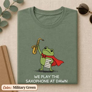 Peut inclure: Un t-shirt vert militaire avec une illustration d'une grenouille verte portant une cape rouge, tenant un saxophone doré et une partition. Sous l'illustration, le texte indique : WE PLAY THE SAXOPHONE AT DAWN. Le t-shirt a un col rond.