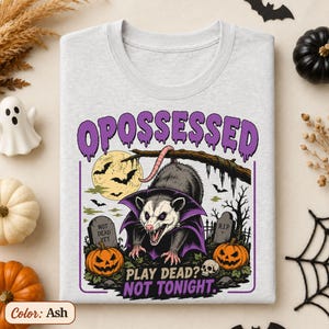 May include: An ash gray t-shirt featuring a graphic of an opossum wearing a purple cape in a graveyard. The text reads OPOSSESSED at the top and PLAY DEAD? NOT TONIGHT. at the bottom, surrounded by pumpkins, tombstones, a full moon, and bats.
