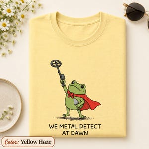 Puede incluir: Una camiseta de color amarillo haze con una ilustración de una rana verde con capa roja, que sostiene un detector de metales y una pala pequeña. Debajo de la rana, el texto WE METAL DETECT AT DAWN aparece escrito en letras mayúsculas negras.
