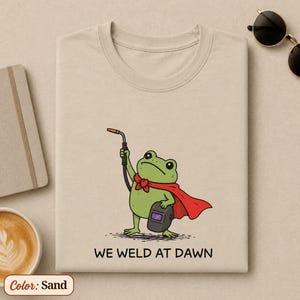 May include: Sand-colored t-shirt featuring a cartoon frog dressed as a superhero, holding a welding torch and a welding mask. The frog wears a red cape and bow tie. The text "WE WELD AT DAWN" is printed below the frog.