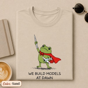 May include: A sand-colored t-shirt featuring a graphic of a green frog wearing a red cape, holding a hobby knife in one hand and a small model airplane in the other. Below the illustration, the text reads: WE BUILD MODELS AT DAWN.
