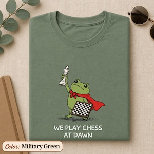 May include: A military green t-shirt featuring a graphic of a green frog wearing a red cape, holding a white chess king piece in one hand and a chessboard in the other. Below the frog, the text reads WE PLAY CHESS AT DAWN in white letters.