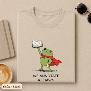 May include: A sand-colored t-shirt featuring a graphic of a green frog wearing a red cape, holding an open book in one hand and a yellow highlighter in the other. Below the frog, the text reads WE ANNOTATE AT DAWN in black capital letters.