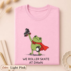 May include: A light pink t-shirt featuring a green frog wearing a red cape and holding a black roller skate. The frog also wears a knee pad on one leg. Below the illustration, the text reads: WE ROLLER SKATE AT DAWN. The design is centered.