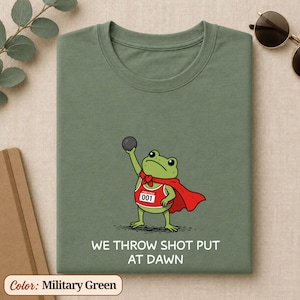 May include: A military green t-shirt featuring a cartoon frog wearing a red cape and a red athletic tank top with the number 001, holding a shot put ball. Below the frog, the text reads WE THROW SHOT PUT AT DAWN in white capital letters.