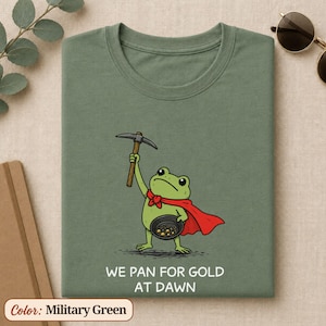 Peut inclure: Un t-shirt vert militaire avec une illustration d'une grenouille verte portant une cape rouge, tenant une pioche et une batée. Sous le dessin, le texte indique : WE PAN FOR GOLD AT DAWN. Le motif est centré sur le devant du vêtement.