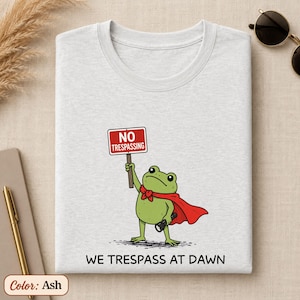 Puede incluir: Una camiseta color ceniza con el gráfico de una rana verde con capa roja, sosteniendo una linterna y un letrero rojo que dice NO TRESPASSING. Debajo de la rana, el texto WE TRESPASS AT DAWN aparece escrito en letras negras y mayúsculas.