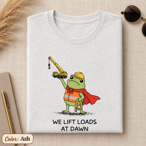May include: An ash-colored t-shirt featuring a graphic of a green frog wearing a yellow hard hat, an orange safety vest, and a red cape. The frog holds a yellow crane above its head. Below the graphic, the text reads: WE LIFT LOADS AT DAWN.