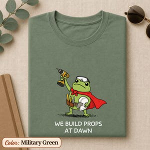 May include: A military green t-shirt featuring a graphic of a green frog wearing a red cape, a tool belt, and safety goggles. The frog holds a yellow power drill in one hand and a white helmet in the other. The text reads WE BUILD PROPS AT DAWN.