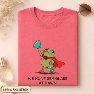 Peut inclure: Un t-shirt couleur corail soie avec une illustration de grenouille verte portant une cape rouge, tenant un morceau de verre de mer turquoise et un filet rempli de verre de mer. Le texte indique : WE HUNT SEA GLASS AT DAWN.